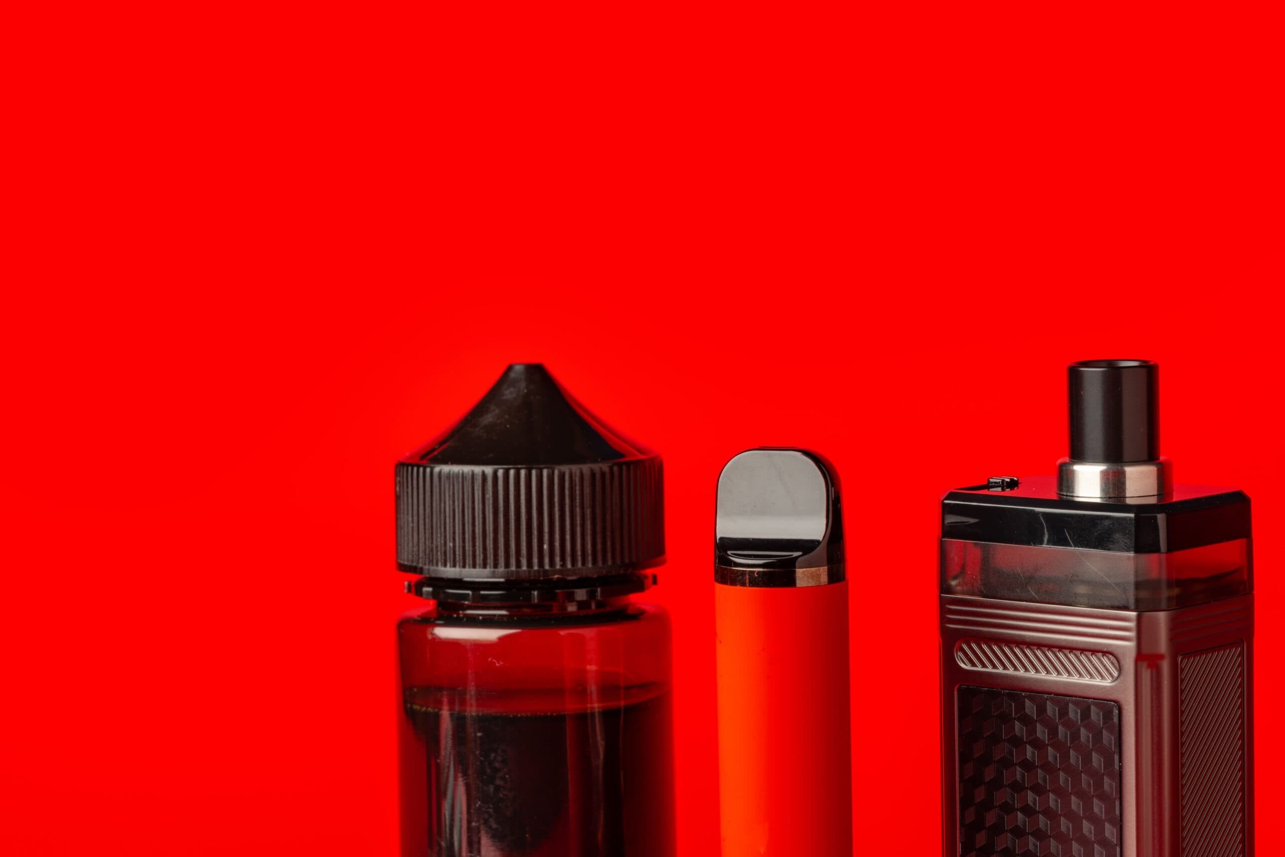 Three vaping products including an e-liquid bottle, a red disposable vape pen, and a refillable vape mod against a vibrant red background.