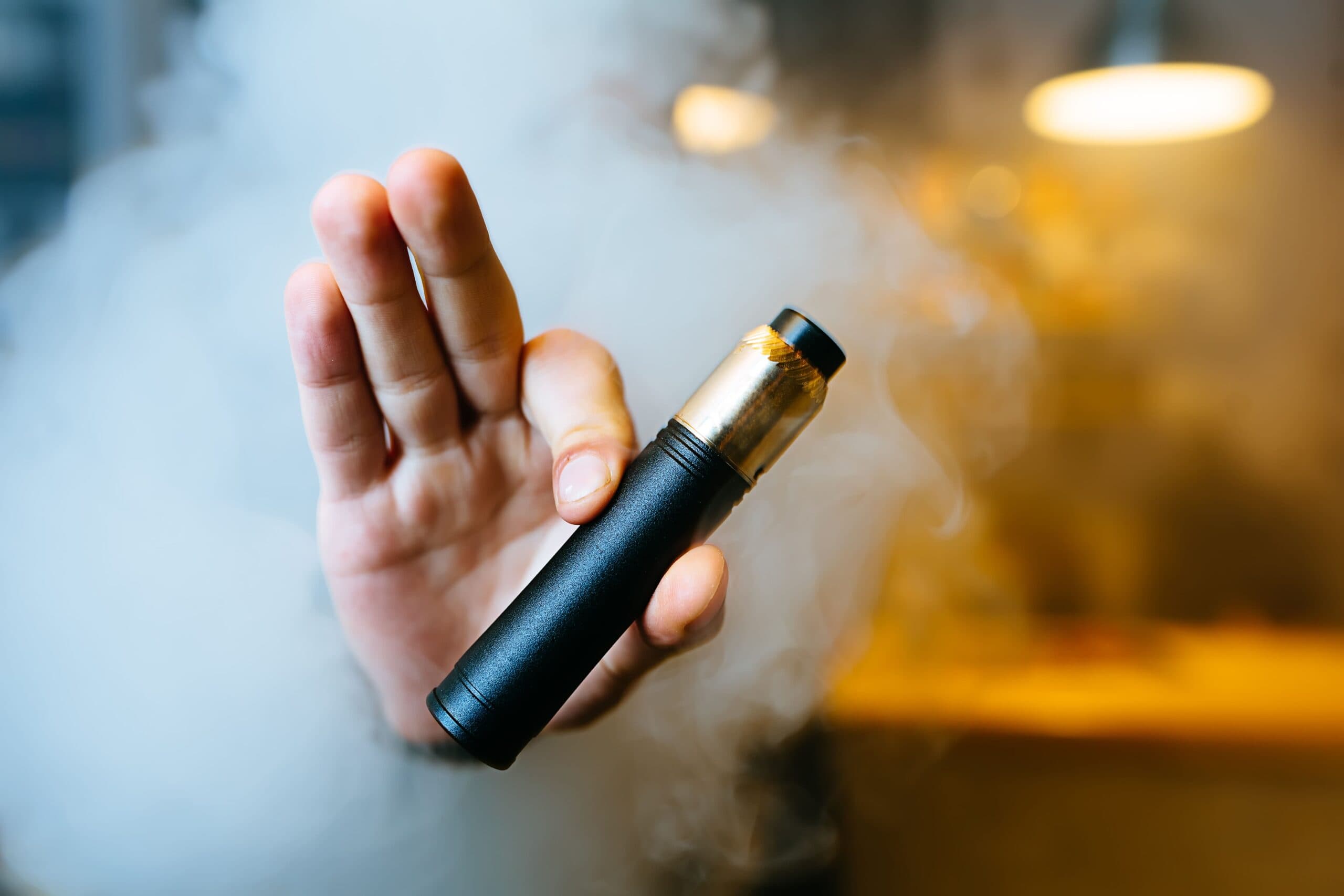 Hand holding a herbal vapouriser surrounded by vapour in a warm indoor setting