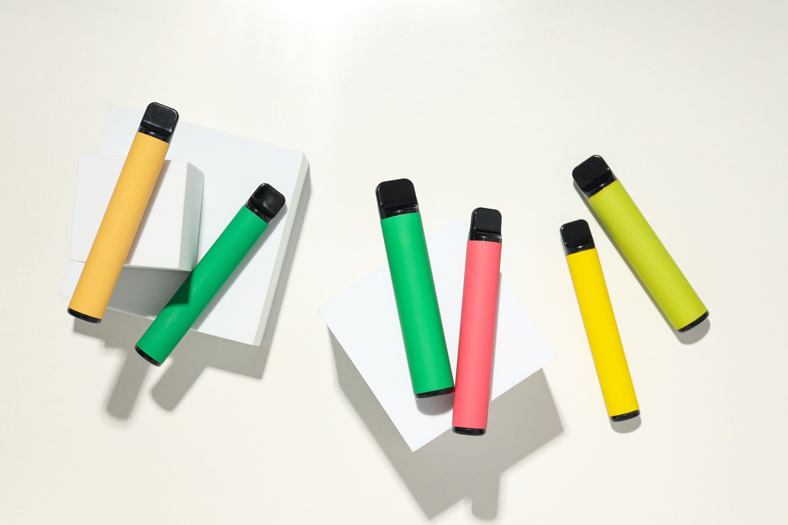 Colorful disposable vape pens in yellow, green, pink, and lime green arranged on white blocks - guide to how long disposable vapes last