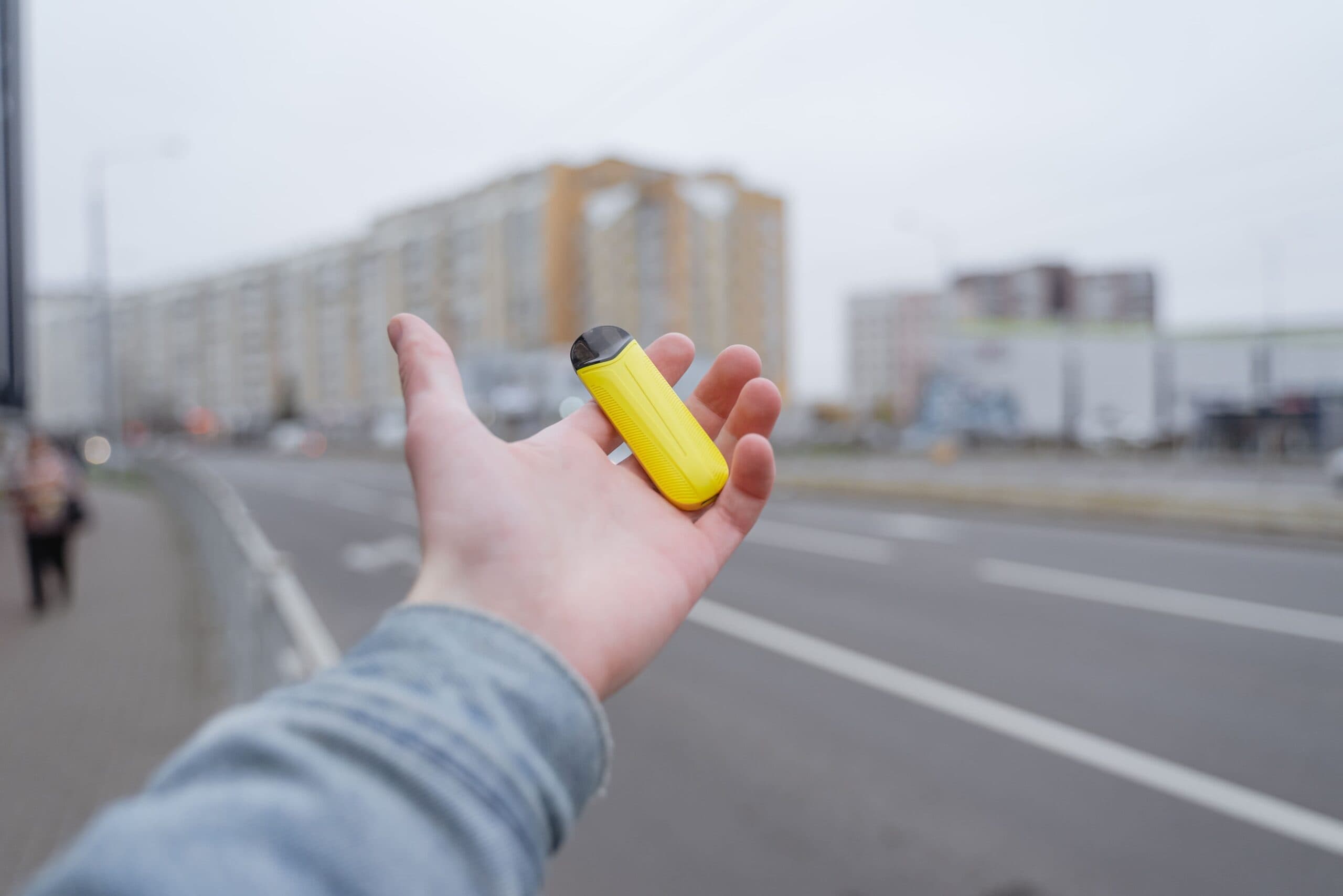 Person holding a compact yellow vape pod system in hand on a city street.